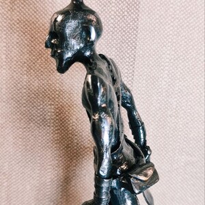 Pu&ograve; includere: Una scultura metallica scura di una figura umanoide con un'acconciatura a chignon, un profilo facciale affilato e una postura curva. La figura indossa polsini e una piccola borsa in vita, realizzata con una finitura riflettente e testurizzata.