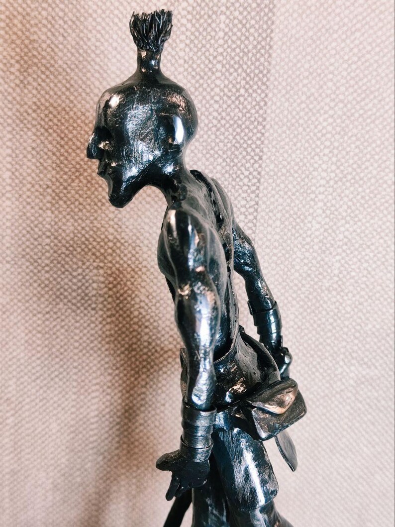 Pu&ograve; includere: Una scultura metallica scura di una figura umanoide con un'acconciatura a chignon, un profilo facciale affilato e una postura curva. La figura indossa polsini e una piccola borsa in vita, realizzata con una finitura riflettente e testurizzata.