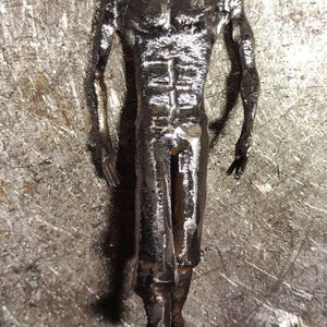 Pu&ograve; includere: Una statuetta metallica color argento che raffigura una forma umana con busto definito, braccia muscolose e gambe. La superficie della scultura presenta una finitura ruvida e strutturata con segni di saldatura visibili.