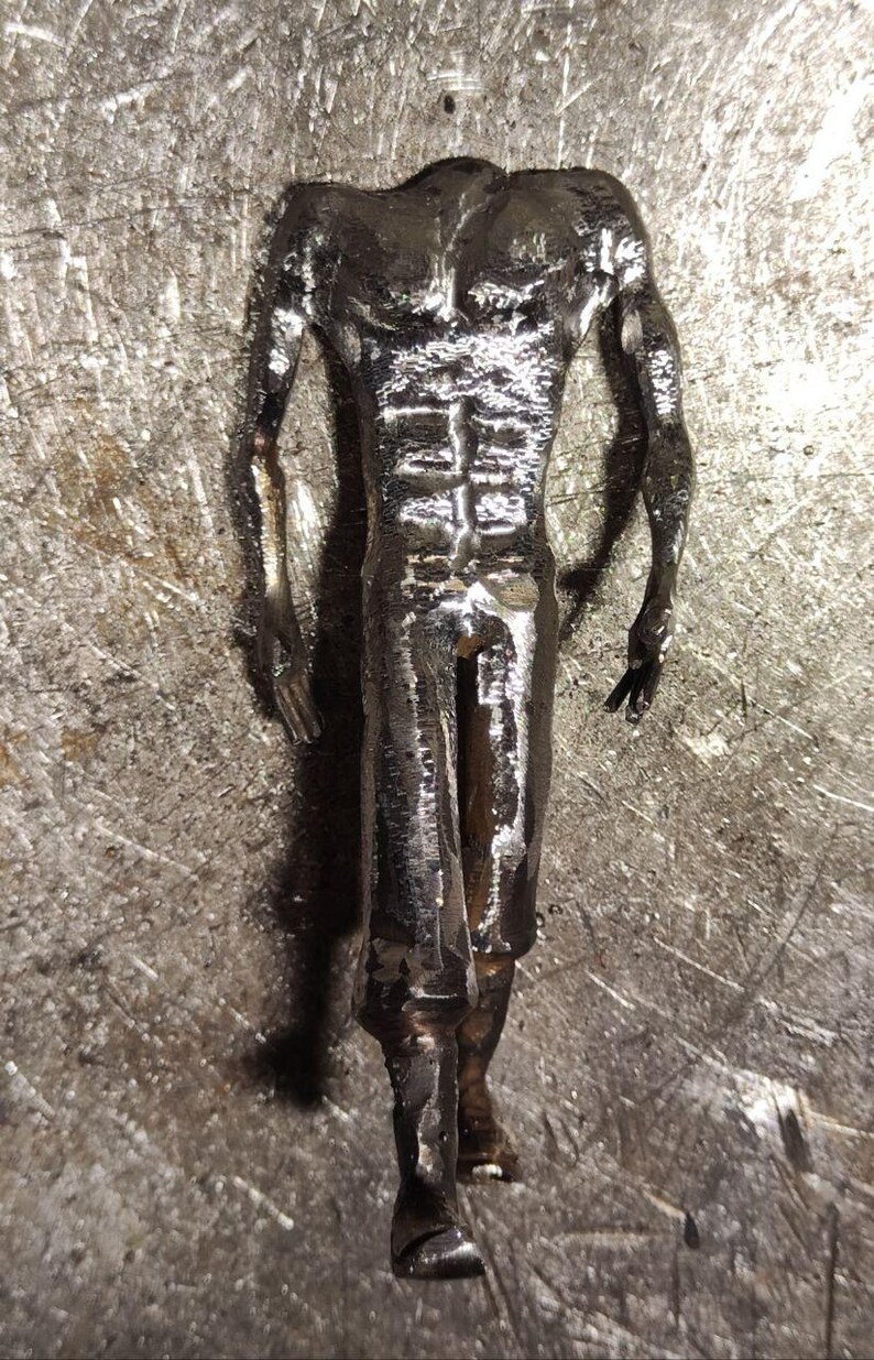 Pu&ograve; includere: Una statuetta metallica color argento che raffigura una forma umana con busto definito, braccia muscolose e gambe. La superficie della scultura presenta una finitura ruvida e strutturata con segni di saldatura visibili.