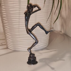 May include: A metal sculpture of a person in a dancing pose. The figure is silver and black, with arms raised and one leg extended. The sculpture is mounted on a base made of stacked metal nuts and bolts. The background includes a white pot and green plant.