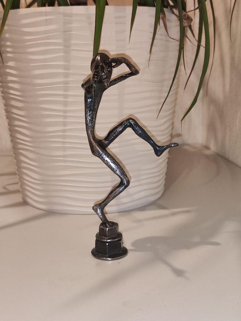 May include: A metal sculpture of a person in a dancing pose. The figure is silver and black, with arms raised and one leg extended. The sculpture is mounted on a base made of stacked metal nuts and bolts. The background includes a white pot and green plant.
