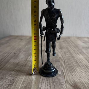 Pu&ograve; includere: Una statuetta metallica scura raffigurante un pirata su una base circolare, che impugna una spada e indossa una cintura con fibbia. Un metro a nastro giallo accanto alla figura indica un'altezza totale di circa 21 cm.