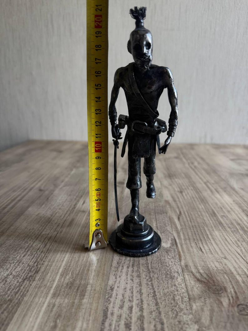 Pu&ograve; includere: Una statuetta metallica scura raffigurante un pirata su una base circolare, che impugna una spada e indossa una cintura con fibbia. Un metro a nastro giallo accanto alla figura indica un'altezza totale di circa 21 cm.