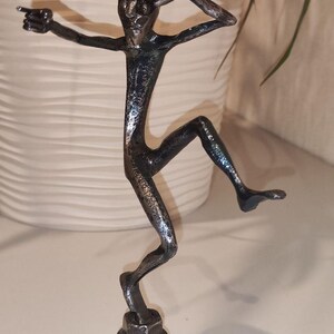 May include: A silver-colored metal sculpture of a stylized figure in a dynamic pose, with one arm outstretched and the other shielding its eyes. The figure is mounted on a hexagonal base, suggesting a modern art piece or decorative item.