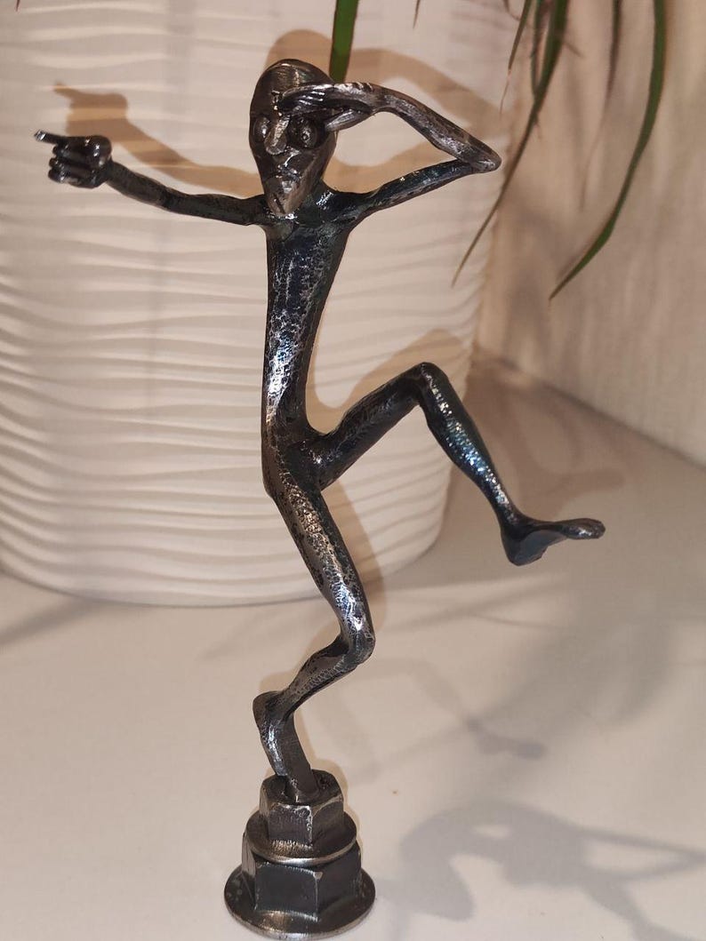 May include: A silver-colored metal sculpture of a stylized figure in a dynamic pose, with one arm outstretched and the other shielding its eyes. The figure is mounted on a hexagonal base, suggesting a modern art piece or decorative item.