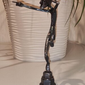 May include: A handcrafted metal sculpture of a slender figure pointing with one arm outstretched. The dark gray sculpture is mounted on a base made of stacked metal nuts. The figure has a textured surface and is set against a neutral background.