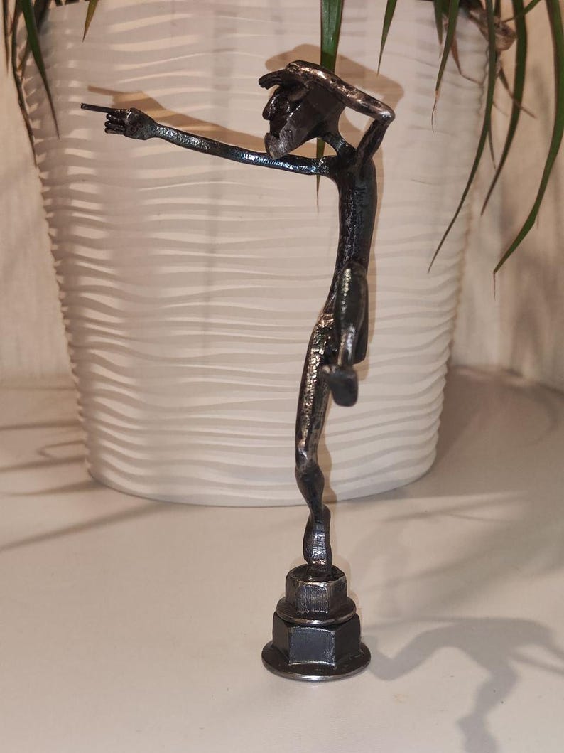 May include: A handcrafted metal sculpture of a slender figure pointing with one arm outstretched. The dark gray sculpture is mounted on a base made of stacked metal nuts. The figure has a textured surface and is set against a neutral background.