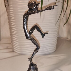 May include: A metal sculpture of a skeletal figure in a dynamic pose, pointing forward. The sculpture is mounted on a hexagonal base. The figure is dark gray and appears to be made of welded metal.