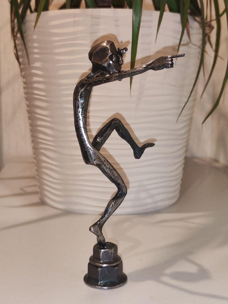 May include: A metal sculpture of a skeletal figure in a dynamic pose, pointing forward. The sculpture is mounted on a hexagonal base. The figure is dark gray and appears to be made of welded metal.