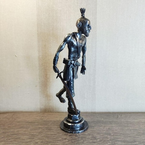 Pu&ograve; includere: Una statuetta metallica color argento raffigurante un personaggio snello e calvo con un nodo tra i capelli, che indossa una cintura e una tunica. La figura poggia su una base circolare e impugna una spada lunga e sottile rivolta verso il basso.