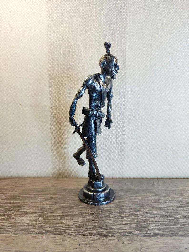 Pu&ograve; includere: Una statuetta metallica color argento raffigurante un personaggio snello e calvo con un nodo tra i capelli, che indossa una cintura e una tunica. La figura poggia su una base circolare e impugna una spada lunga e sottile rivolta verso il basso.