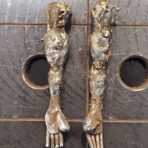 Pu&ograve; includere: Due sculture di braccia in metallo con una finitura ruvida e testurizzata e segni di saldatura visibili. Le braccia sono posizionate verticalmente, mostrando strutture delle mani dettagliate con dita e pollice distinti in toni argento e bronzo.