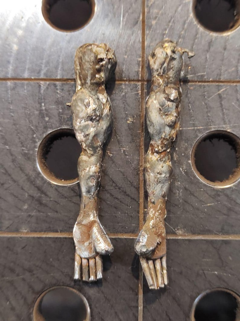 Pu&ograve; includere: Due sculture di braccia in metallo con una finitura ruvida e testurizzata e segni di saldatura visibili. Le braccia sono posizionate verticalmente, mostrando strutture delle mani dettagliate con dita e pollice distinti in toni argento e bronzo.