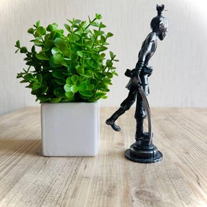 Pu&ograve; includere: Una statuetta color grigio scuro metallizzato raffigurante un personaggio slanciato e stilizzato in piedi su una base circolare, che impugna un oggetto ricurvo simile a una spada. La figura ha un chignon ed &egrave; accanto a una piccola pianta artificiale verde.