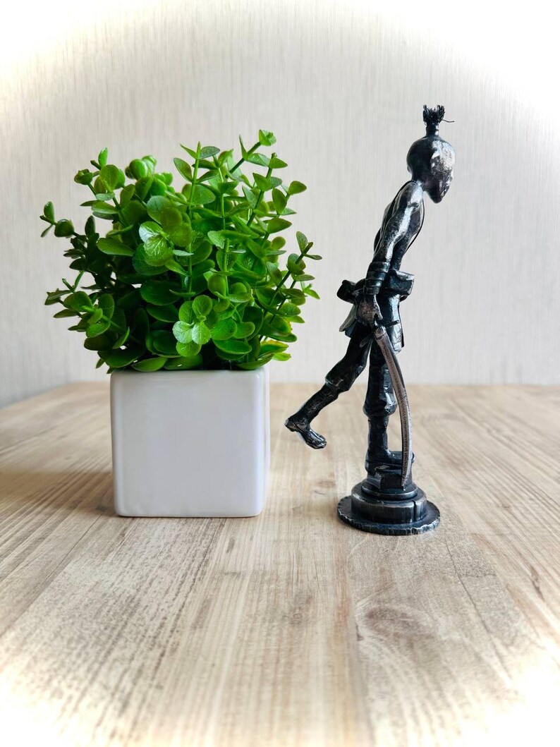 Pu&ograve; includere: Una statuetta color grigio scuro metallizzato raffigurante un personaggio slanciato e stilizzato in piedi su una base circolare, che impugna un oggetto ricurvo simile a una spada. La figura ha un chignon ed &egrave; accanto a una piccola pianta artificiale verde.
