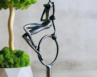 May include: A silver-toned metal sculpture of a stylised figure sitting on a circular hoop mounted on a pedestal base. The figure has an elongated form with a smooth, reflective surface. Beside it stands a small tree with bright green moss foliage.