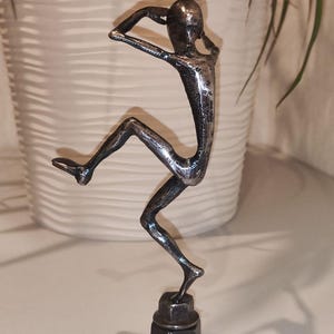 May include: A silver metal sculpture of a person in a dynamic pose, with arms raised and one leg extended. The figure is mounted on a base made of stacked metal nuts. The sculpture has a textured surface and is likely handmade.