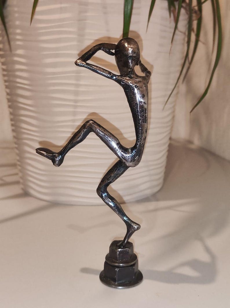 May include: A silver metal sculpture of a person in a dynamic pose, with arms raised and one leg extended. The figure is mounted on a base made of stacked metal nuts. The sculpture has a textured surface and is likely handmade.