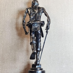 Pu&ograve; includere: Una scultura metallica scura di una figura in piedi vista da dietro. La figura ha un'acconciatura a crocchia, indossa una cinghia sulla schiena e tiene in mano un oggetto sottile a forma di asta. La scultura &egrave; montata su una base circolare.