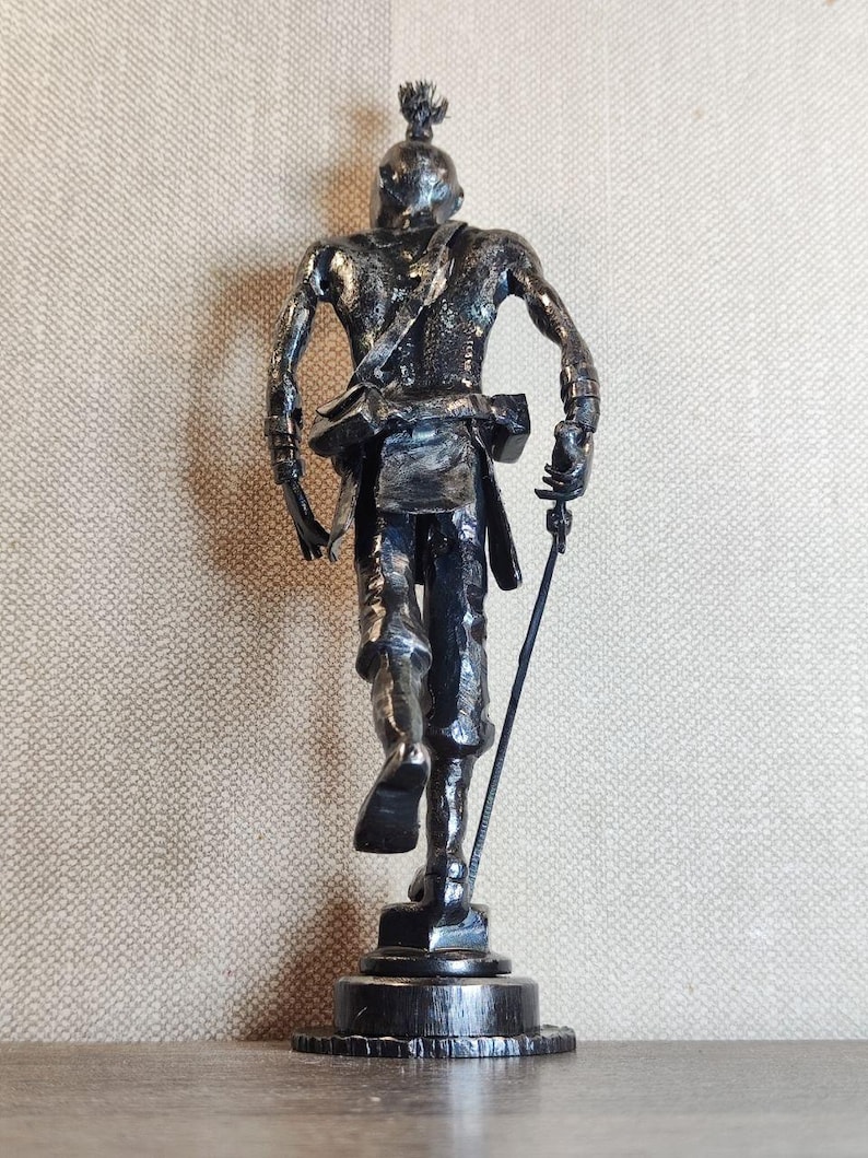 Pu&ograve; includere: Una scultura metallica scura di una figura in piedi vista da dietro. La figura ha un'acconciatura a crocchia, indossa una cinghia sulla schiena e tiene in mano un oggetto sottile a forma di asta. La scultura &egrave; montata su una base circolare.