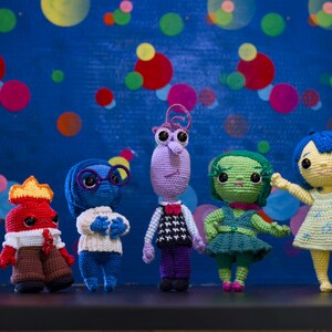 Puede incluir: Un conjunto de siete juguetes de peluche tejidos a crochet inspirados en los personajes de la película Inside Out. Los juguetes son de diferentes colores y tienen diferentes expresiones faciales. Están de pie en fila sobre un fondo azul con lunares blancos.