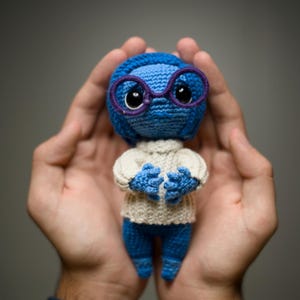 Puede incluir: Un personaje de crochet azul con gafas moradas y un suéter blanco. El personaje tiene una expresión triste y está siendo sostenido en las manos de alguien.