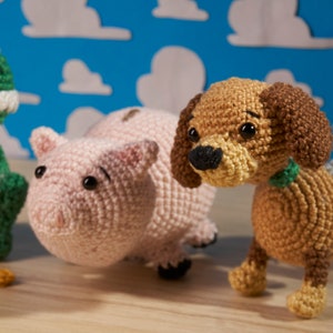Puede incluir: Tres juguetes de peluche de crochet: un dinosaurio verde, un cerdo rosa y un perro marrón con una cola rayada gris y blanca.