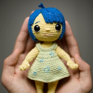 Puede incluir: Una muñeca de crochet con pelo azul, piel amarilla y un vestido verde. La muñeca sostiene una pequeña flor azul en su mano.
