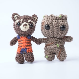 Puede incluir: Dos juguetes de peluche tejidos a crochet, uno marrón y naranja con una cara de mapache y el otro marrón con una cara de árbol.