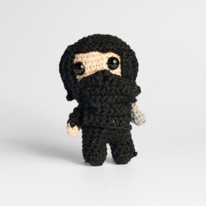 May include: A crocheted plush toy of a superhero character wearing a black mask and costume. The toy has black hair, a beige face, and large black eyes.