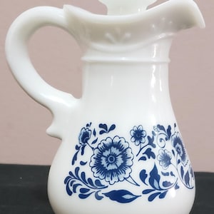 Könnte beinhalten: Weißer Keramik-Sahnekrug mit Henkel und Ausguss, verziert mit einem blauen Blumenmuster. Der Krug hat einen Deckel mit einem kleinen Knauf. Das Design umfasst detaillierte blaue Blumen und Blätter, was ihm ein klassisches, dekoratives Aussehen verleiht.