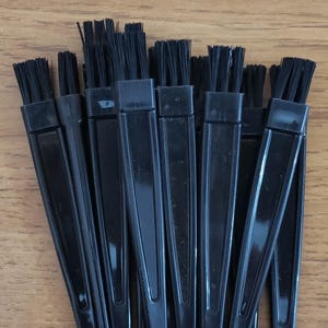 May include: A collection of black handled brushes with black bristles. The brushes have a triangular handle and are arranged in a fan-like pattern. The bristles are tightly packed and appear to be made of synthetic fibers. The brushes are likely for applying makeup or other cosmetic products.