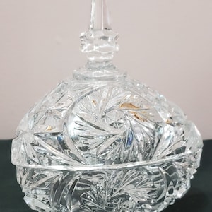 May include: A clear, cut-glass candy dish with a lid and three small feet. The dish features a detailed, starburst pattern across its surface, with a pointed finial on the lid. The design catches and reflects light, creating a sparkling effect.