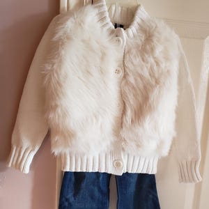 May include: A white knit cardigan with a faux fur front panel, button closure, and long sleeves. Paired with dark blue denim flared jeans. The outfit is displayed on a hanger.