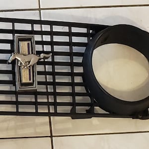 May include: Black car grille with a rectangular grid pattern and two circular headlight openings. A silver and gold emblem of a running horse is centered on the grille.