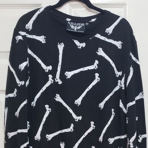 May include: A black long-sleeve shirt featuring an all-over white bone print pattern. The shirt has a crew neckline, a high-low hemline, and a Killstar brand label at the neck and bottom hem. The fabric is black with white skeletal bone graphics.