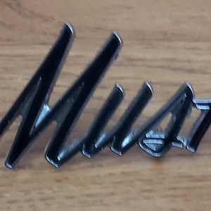 Puede incluir: Un emblema "Mustang" cromado y negro, probablemente para un coche, se muestra sobre un fondo de vetas de madera. La fuente cursiva y la superficie reflectante del emblema sugieren una estética clásica o vintage. La palabra "Mustang" es claramente visible.