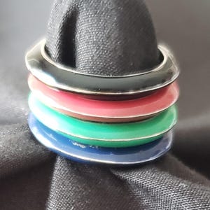 May include: A set of four colourful rings, in black, red, green, and blue. The rings have a shiny, metallic finish and are stacked. The rings are set against a dark background.