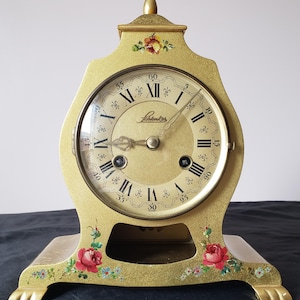 May include: A gold-colored, vintage mantel clock with a floral design. The clock face has Roman numerals and the brand name "Lahrkes". The clock is decorated with red and blue flowers.