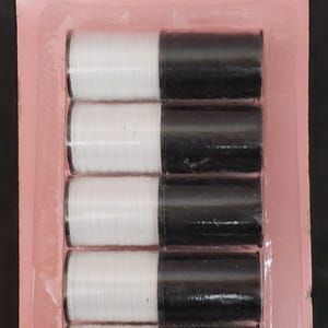 Peut inclure: Un paquet de 12 bobines de fil à coudre, totalisant 32 mètres, présenté sur un support en carton rose avec des rayures rouges et blanches. L'ensemble contient six bobines de fil blanc et six bobines de fil noir, scellées sous plastique.