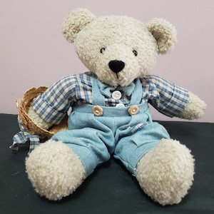 May include: A cream-colored teddy bear with textured fur, wearing a blue and white plaid long-sleeve shirt under light blue denim overalls with two white buttons at the waist and a small straw hat tucked under one arm with a plaid ribbon.