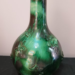 Può includere: Un vaso in ceramica smaltata verde con corpo bulboso e collo alto e sottile. Il vaso presenta motivi floreali e a stella marina in rilievo. La smaltatura ha una finitura lucida con sfumature marroni e verdi. Il vaso è alto circa 30 cm.