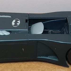 May include: A dark grey toy Batmobile vehicle featuring a sleek, elongated body, a silver Batman logo on the bonnet, light grey side vents, a light grey open cockpit seat, and silver-coloured wheels with a pointed, fin-like rear design.