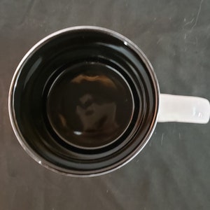 May include: Overhead view of a white ceramic mug with a black interior. The mug has a white handle and a shiny, reflective surface. The mug is empty and sits on a dark gray surface.