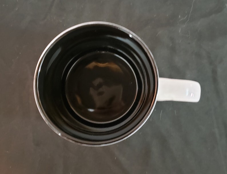 May include: Overhead view of a white ceramic mug with a black interior. The mug has a white handle and a shiny, reflective surface. The mug is empty and sits on a dark gray surface.