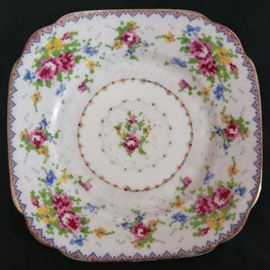 May include: Square white plate with a floral design. The plate features a pattern of pink, yellow, and blue flowers arranged in a pixelated style. The rim has a gold trim and a blue and pink border.