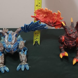 May include: Two toy dragon figures. One is blue and silver, the other is red and black. Both have large wings and are posed on all fours. A yellow measuring tape is between them.