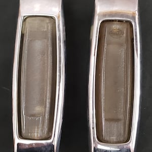 May include: Two vintage rectangular automotive lights with chrome bezels. Each light has a clear, ribbed lens and a silver frame. The lights are likely for a classic car or truck.