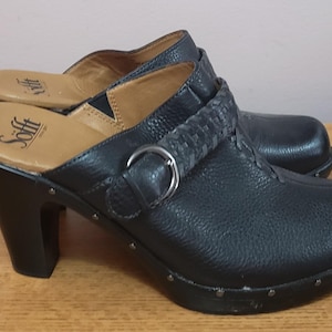 May include: A pair of black leather clogs with a textured finish, featuring a braided strap across the top, a silver-tone metal buckle, and a block heel. The interior footbed is tan and displays the Sofft brand logo in black lettering.